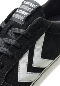 Czarne zamszowe sneakersy z białym skórzanym wzorem w kształcie chevronu, wyposażone w czarne sznurówki i etykietę z logo na języku. Widoczna gumowa podeszwa.