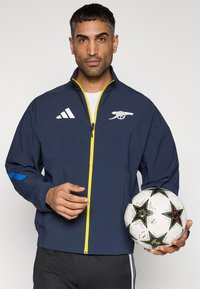Αθλητικό μπουφάν σε ναυτικό μπλε με κίτρινο φερμουάρ και μπλε λεπτομέριες, με λογότυπο στο στήθος, συνοδευόμενο από ποδοσφαιρική μπάλα σε μαύρο και λευκό μοτίβο.