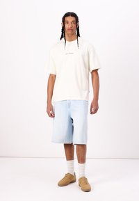 Light cream oversized t-shirt with embroidered text, paired with light blue denim shorts. Footwear: tan sneakers, white ankle socks.