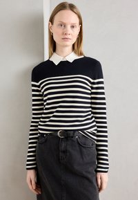 Tengerés sötétkék és krémszínű csíkos pulóver fehér gallérral, párosítva egy fekete farmer szoknyával és egy fekete övvel, ezüst csattal.