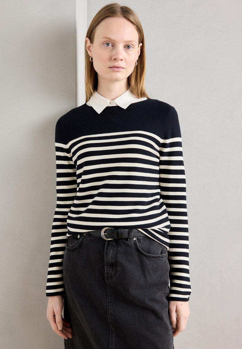 Tengerés sötétkék és krémszínű csíkos pulóver fehér gallérral, párosítva egy fekete farmer szoknyával és egy fekete övvel, ezüst csattal.