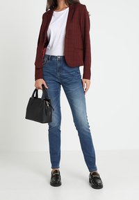 Žena v vínovém saku, bílém tričku, modrých džínách a černých mokasínech, držící černou kabelku na prostém bílém pozadí.