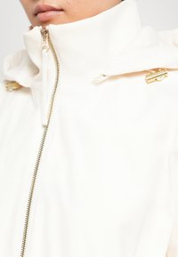 Witte jas met een hoge kraag, gouden hardware accenten en een zichtbare ritssluiting. Gladde textuur met een modern ontwerp.