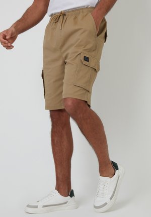 Pantalones cortos de color arena con cintura elástica, que cuentan con bolsillos laterales y traseros, y una tela texturizada. Combinados con zapatillas blancas y detalles en verde.