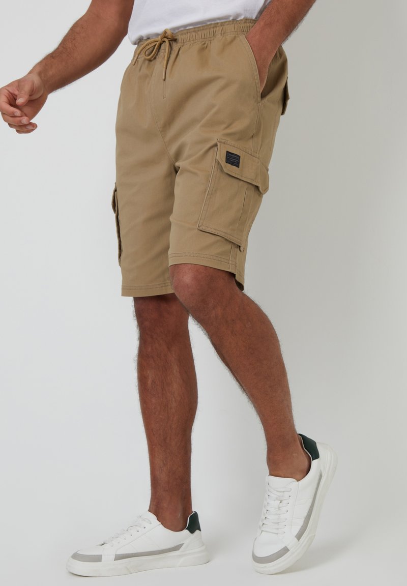 Tan cargo shorts med elastisk talje, med sidelommer og baglommer, samt en tekstureret stof. Sammen med hvide sneakers og grønne accenter.