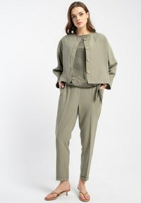 Oliwkowa kurtka z dużymi guzikami, nałożona na wzorzysty top, w zestawieniu z pasującymi spodniami. Gładka tekstura, luźny krój, casualowy styl.