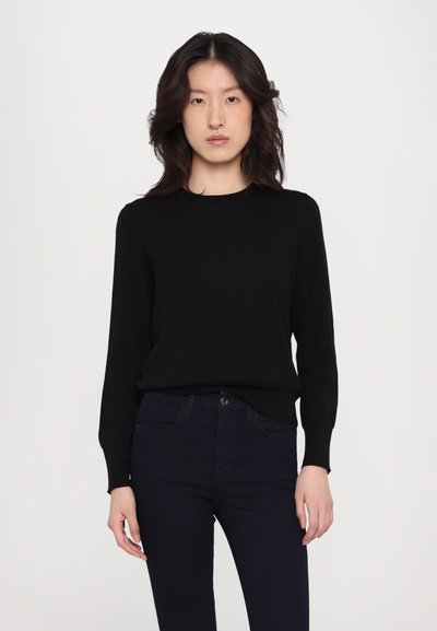 Femeie cu păr dark, până la umeri, îmbrăcată cu un top simplu negru cu mânecă lungă și pantaloni închisă, stând pe un fundal deschis.