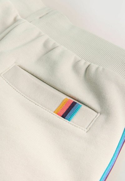 Flødefarvet stof med elastisk taljebånd og en lomme med en lodret stribe i orange, pink, lilla og blå.