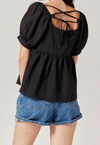 Black puff-sleeve top with a peplum design, featuring a crisscross back tie and gathered waist. Paired with blue denim shorts.