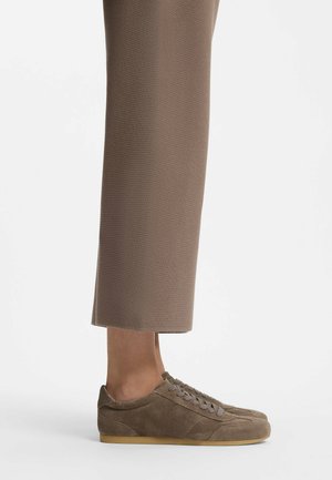 Personne portant des baskets en daim marron à lacets avec des semelles en gomme et un pantalon marron raccourci sur un fond blanc uni.
