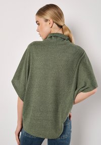 Pull vert à côtes avec un col haut et des manches courtes. Le dos présente un ourlet légèrement plongeant et une coupe décontractée. Porté avec un jean en denim.