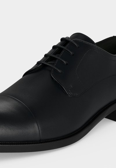Chaussure habillée en cuir noir avec bout droit et laçage fermé sur une semelle lisse sur fond gris.