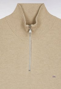 Gilet zippé beige avec un col montant. Dispose d'un tissu en tricot texturé et d'un petit logo sur le coin inférieur droit. Détail de fermeture éclair argentée.