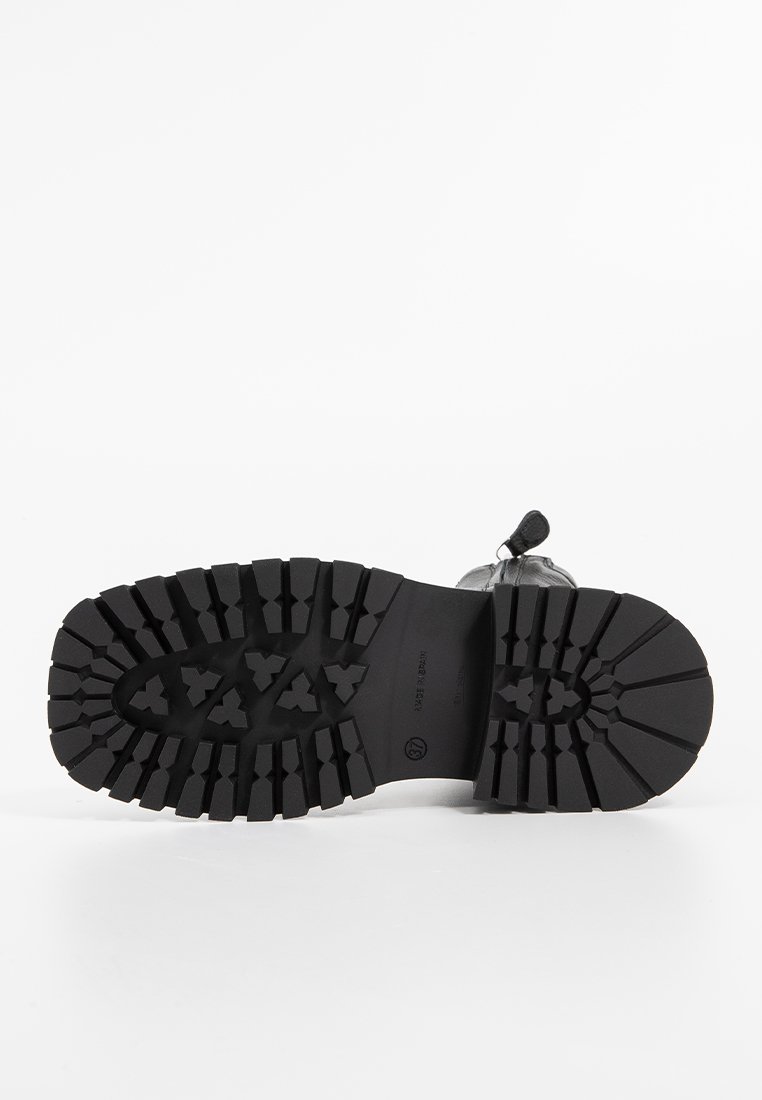Semelle de botte en caoutchouc noir avec des crampons profonds et robustes ainsi qu'une surface texturée. Présente une empreinte de logo près du talon pour un détail supplémentaire.