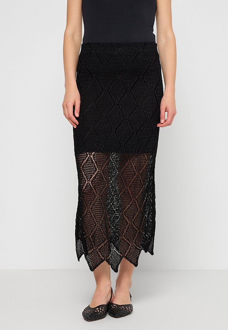 Frau in einem schwarzen ärmellosen Oberteil und einem schwarzen gehäkelten Midirock mit Diamantmuster, kombiniert mit schwarzen geflochtenen flachen Schuhen.