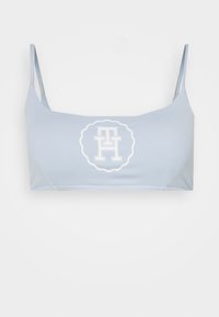 Světle modrý crop top s tenkými ramínky, s bílým kruhovým logem ve středu, na kterém jsou překrývající se písmena. Jemná, hladká textura látky.
