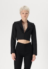 Svart kort blazer med lange ermer, dyp V-hals og frontlukking med knapper. Stylet med høytlivte svarte bukser, noe som skaper en stram silhuett.
