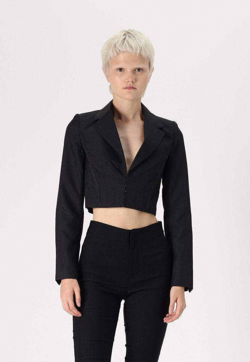 Svart kort blazer med lange ermer, dyp V-hals og frontlukking med knapper. Stylet med høytlivte svarte bukser, noe som skaper en stram silhuett.