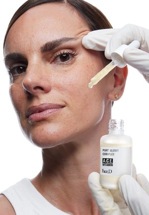 Bottiglia di siero trasparente con un'etichetta bianca, tenuta accanto al viso di una donna. Indossa un top bianco e guanti in lattice, con una carnagione liscia.