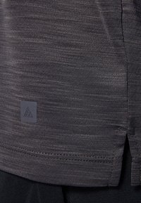 Camisa de tejido técnico gris con un patrón texturizado, aberturas laterales y un pequeño parche de logo cerca del dobladillo. Material suave que absorbe la humedad.