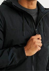 Zwarte lichte jas met capuchon, ritssluiting aan de voorkant en geribbelde manchetten. Heeft een gladde textuur en verstelbare koordjes.