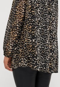 Blouse imprimé léopard en tissu léger et transparent avec des taches noires et beige. Manches longues froncées et détail d'ourlet arrondi.