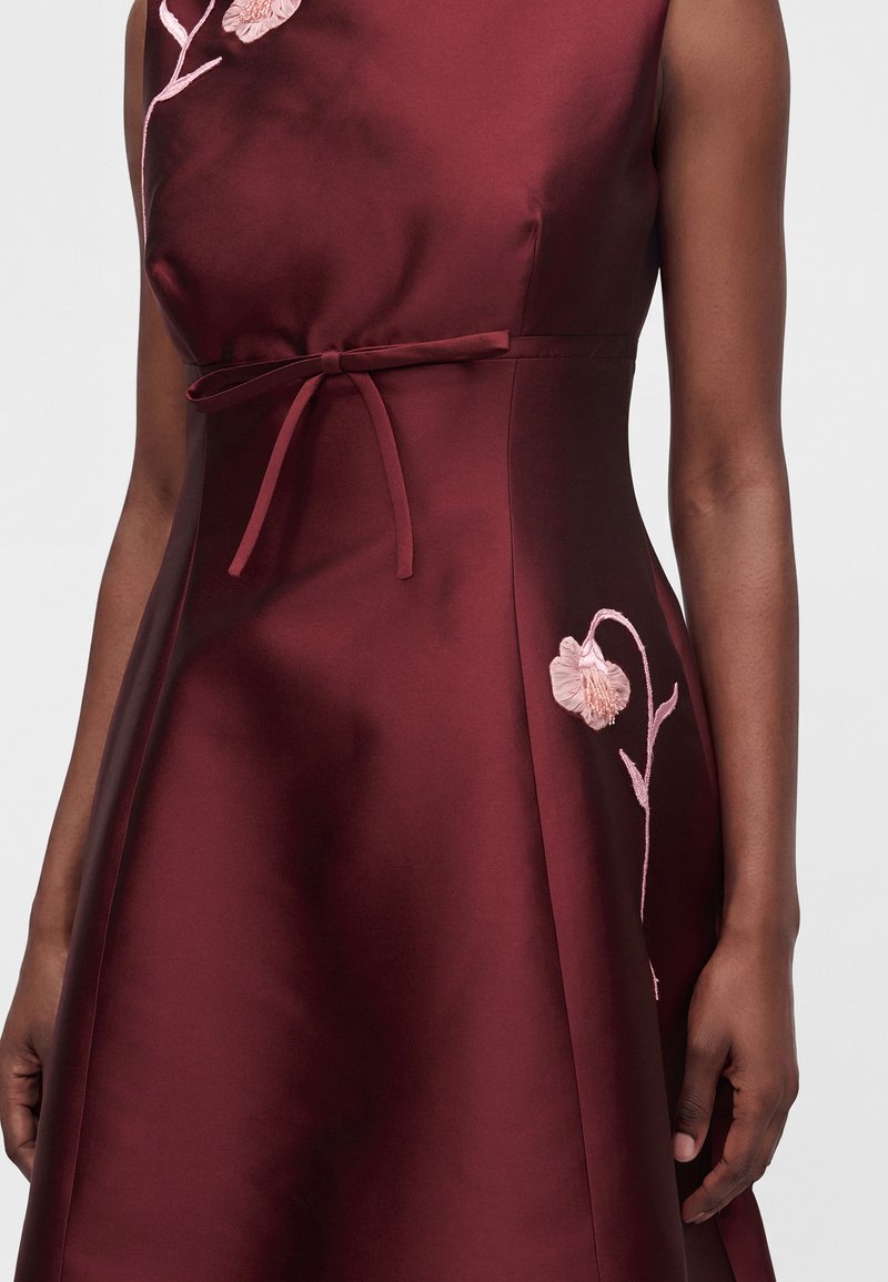 Femme portant une robe en satin bordeaux sans manches, ceinturée à la taille et ornée d’une broderie florale rose sur la poitrine et le bas de la jupe.