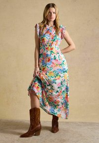 Sieviete valkā krāsainu midi ziedu kleitu ar volāniem piedurknēs un brūnas ādas kaubojubotas, stāvot pret vienkrāsainu bēšu fona sienu.