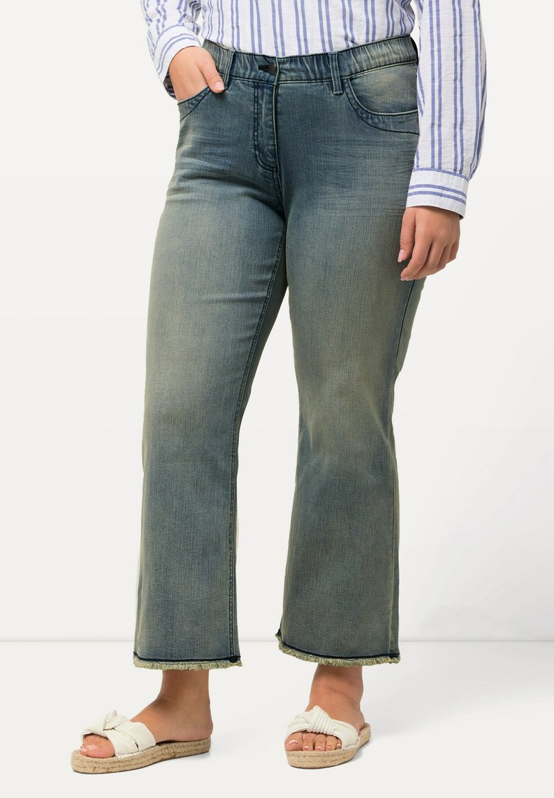 Pantalones vaqueros de mezclilla azules acampanados con un lavado deslavado, diseño de cinco bolsillos y dobladillo deshilachado, combinados con sandalias deslizables blancas con nudos.