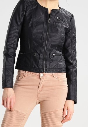 Vrouw die een aangesloten zwart leren jasje met ritssluitingen en lichtroze geribde broek draagt, staand tegen een effen achtergrond.