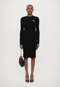 Rochie neagră din tricot cu model rib, cu mâneci lungi, decolteu rotund și un detaliu brodat subtil. Purtați cu sandale negre cu barete și o geantă neagră.