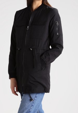 Femme portant une longue veste noire zippée avec des poches sur la poitrine et les manches, une main dans une poche, associée à un jean bleu.