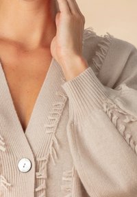 Beige gerippter Strickcardigan mit Knopfverschluss, der Fransenakzente entlang der Vorderseite und eine weiche Textur aufweist, was das kuschelige Design betont.