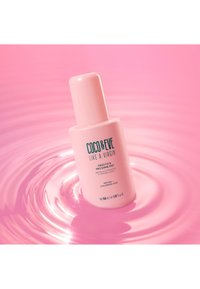Coco & Eve Frizz Fix Pro Shine Mist i en rosa flaska, cylindrisk form, med mörkblå text och ett rundat lock, omgiven av mjuka rosa vågor.