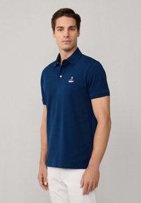 Polo en coton mélangé bleu marine, avec un col à trois boutons et un logo brodé sur la poitrine gauche. Coupe classique, manches courtes.
