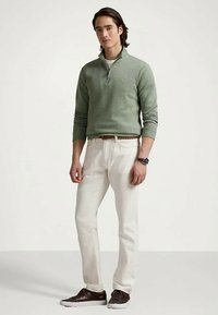 Man med en salviagrön tröja med kvartsyzip, vita byxor med brunt flätat bälte, mörkbruna sneakers och ett armbandsur som står framför en enkel bakgrund.