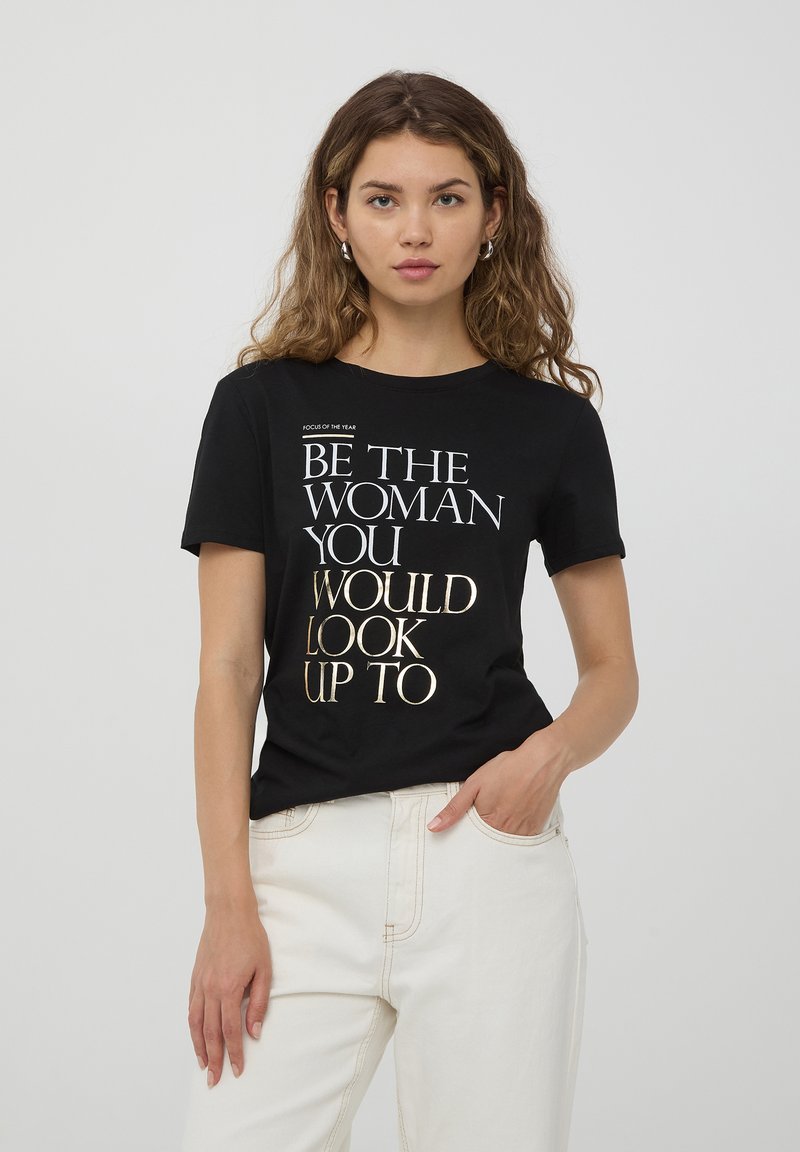 T-shirt in cotone nero con maniche corte, con testo dorato e bianco: "SII LA DONNA A CUI TI PIACEREBBE RIVOLGERTI." Taglio classico, colletto rotondo.