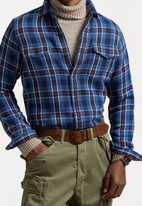 Camicia di flanella a quadri blu con accenti bianchi e rossi, abbinata a un dolcevita beige e pantaloni cargo verde oliva, completata da una cintura marrone.