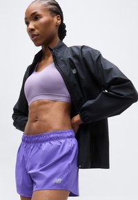 Αθλητική γυναίκα με μωβ αθλητικό σουτιέν και σορτσάκι φοράει μαύρο μπουφάν New Balance, με τα χέρια στη μέση, μπροστά σε απλό φωτεινό φόντο.