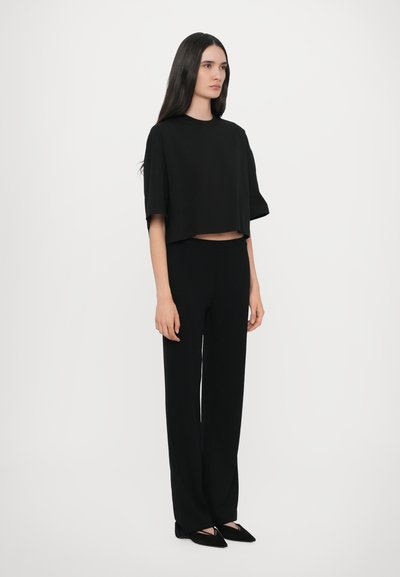 Schwarzes, kurzes Oberteil mit weiten Ärmeln, kombiniert mit schwarzen, geraden Hosen. Weicher Stoff mit glatter Textur, dazu spitze schwarze Schuhe.