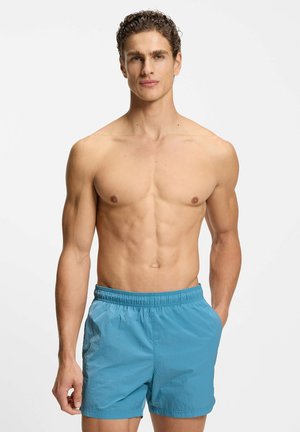 Junger Mann mit kurzen lockigen Haaren, oberkörperfrei, trägt hellblaue Badeshorts vor schlichtem weißem Hintergrund.