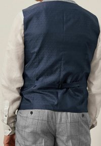 Gilet blu navy con un motivo geometricamente impresso, caratterizzato da una vita con cintura. Indossato sopra una camicia bianca a maniche lunghe e pantaloni grigi a quadretti.
