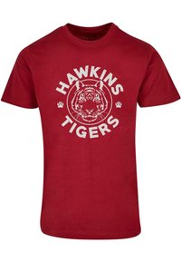 Raudona medvilninė T-marškiniai su apskritimu su tigro veidu, tekstu "HAWKINS TIGERS" ir baltomis letenų pėdomis. Trumpomis rankovėmis ir apvaliu kaklu.