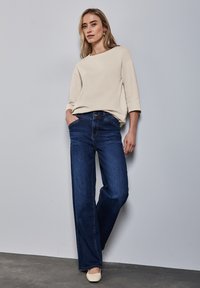 Béžový svetr volného střihu s širokým výstřihem, spárovaný s tmavě modrými, vysokým pasem a širokými kalhotami, a světlými plochými botami.