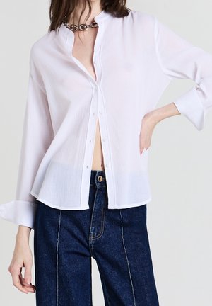 Frau trägt eine locker sitzende weiße Bluse mit Knöpfen und hochgekrempelten Ärmeln sowie dunkelblaue Jeans mit hoher Taille, accessoriert mit einer silbernen Kettenhalskette.