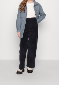 Světle modrá bunda přes bílý strukturovaný top, doplněná o tmavé manšestrové kalhoty s vysokým pasem a širokými nohavicemi, a robustní černé nazouvací boty.