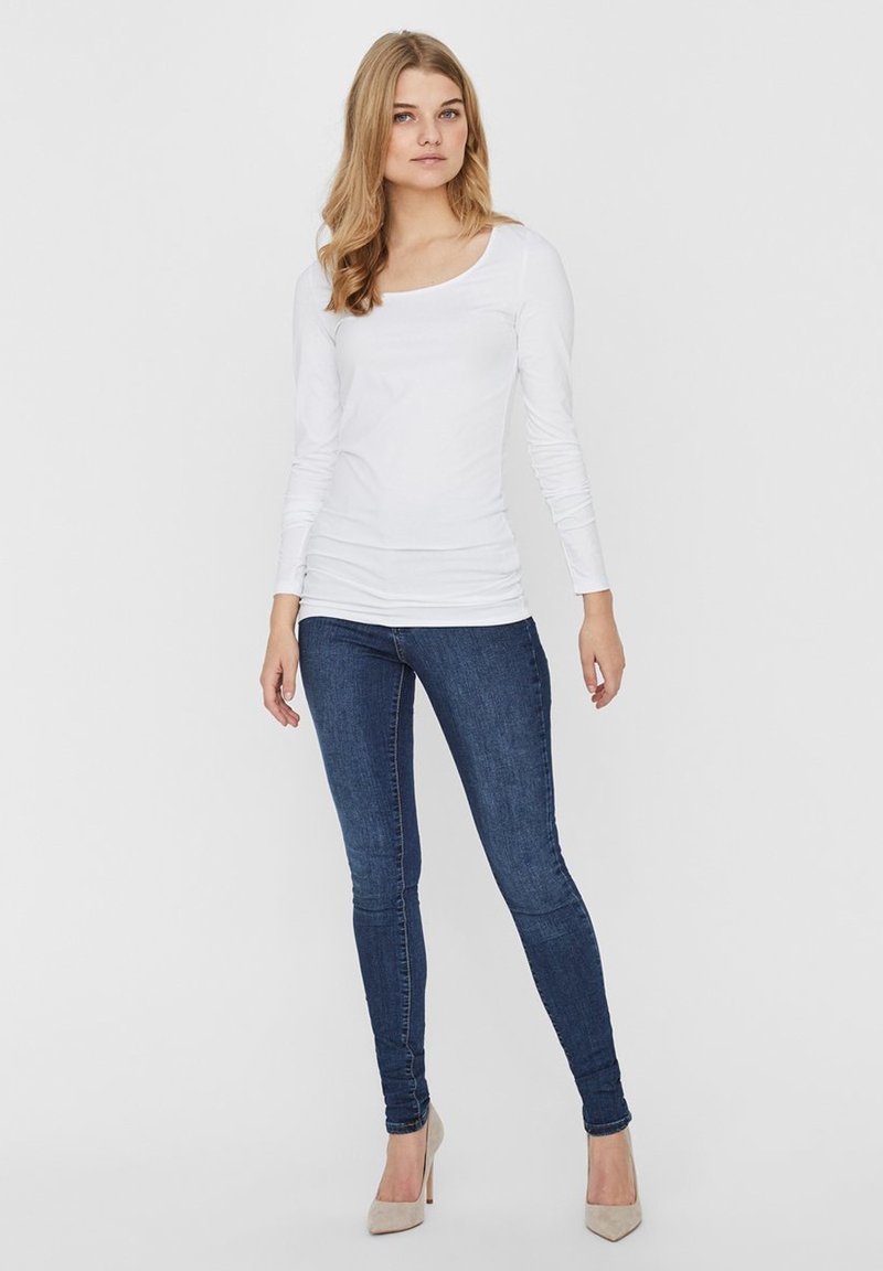 Witte langemouwenshirt met een ronde halslijn, gecombineerd met donkerblauwe skinny jeans en beige hoge hakken, tegen een neutrale achtergrond.