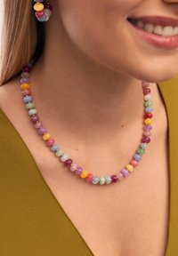 Nahaufnahme einer lächelnden Person, die eine mehrfarbige Perlenkette und passenden Ohrring mit rundlichen Perlen auf einem senffarbenen Oberteil trägt.