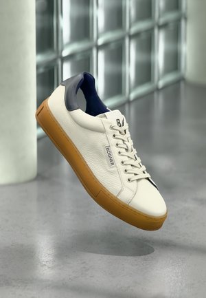 Weißer Ledersneaker mit brauner Sohle und navyblauem Fersendetail, gebrandet mit "Bogner", schwebt über einem reflektierenden grauen Boden.