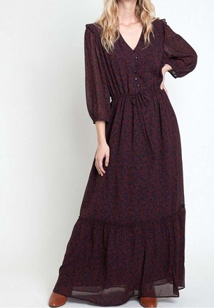 Femme portant une longue robe fleurie violet foncé avec des manches bouffantes, des boutons sur le devant et des bottines marron, debout avec une main sur la hanche.